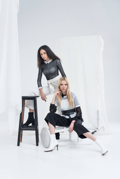 attractive multiethnic women in fashionable black and white clothes posing on chairs near tulle on white - Foto, Imagem