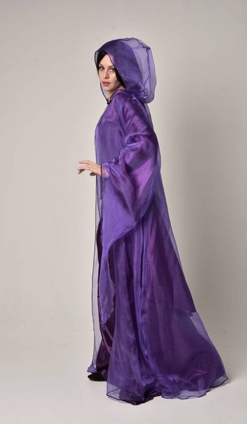 retrato de longitud completa de hermosa chica con el pelo largo y negro, usando vestido medieval de fantasía púrpura y capa. pose de pie sobre fondo gris estudio
. - Foto, Imagen