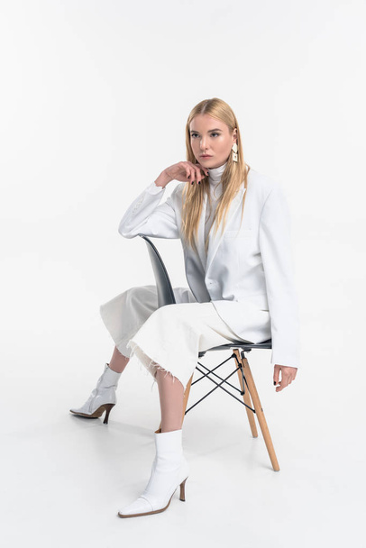 attractive caucasian blonde woman in fashionable white clothes sitting on chair and looking away isolated on white - Foto, Bild