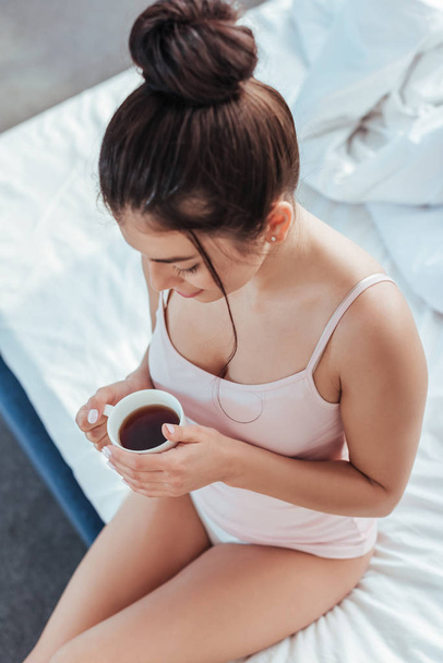 high angle view of young woman sitting with coffee cup on bed during morning time at home - Fotografie, Obrázek