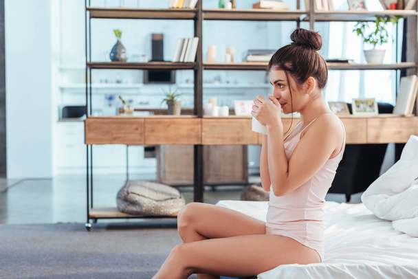 side view of young woman sitting with coffee cup on bed during morning time at home - Фото, изображение