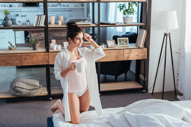 hoge hoekmening van meisje in wit overhemd koffiekopje houden tijdens ochtend tijd thuis - Foto, afbeelding