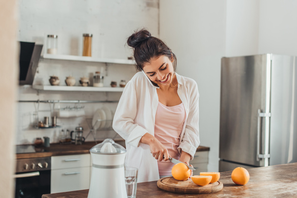 laughing young woman cutting orange by knife and talking on smartphone in kitchen at home - Photo, image