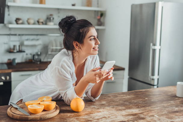 allegra giovane donna utilizzando smartphone a tavolo in legno con arance in cucina a casa
 - Foto, immagini