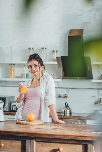 selective focus of young woman holding cup with orange juice in kitchen during morning time at home - Fotoğraf, Görsel