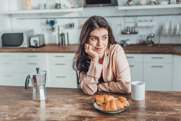 thoughtful young woman sitting at wooden table with coffee cup and croissants in kitchen at home - Φωτογραφία, εικόνα