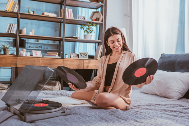 smiling girl holding vinyl record near phonograph and sitting on bed at home - Photo, Image