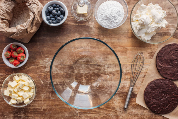 top view of empty glass bowl, whisk and ingredients for delicious homemade cake on wooden table - Foto, Imagem