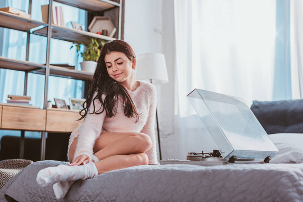 atraktivní usměvavá dívka sedící na posteli a poslech audio přehrávač vinyl doma - Fotografie, Obrázek