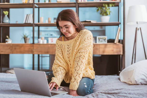 alegre freelancer femenino en gafas que trabajan en el ordenador portátil mientras está sentado en la cama en casa
 - Foto, imagen