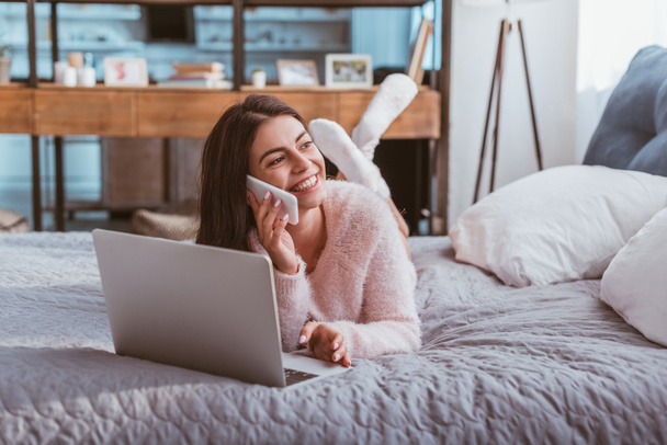 lachende junge Freiberuflerin im Smartphone-Gespräch, während sie zu Hause mit Laptop im Bett liegt - Foto, Bild