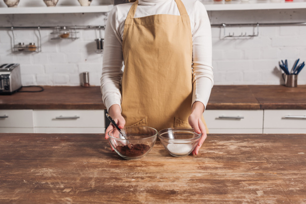 cropped shot of woman in apron holding glass bowls with ingredients for delicious homemade cake - Valokuva, kuva