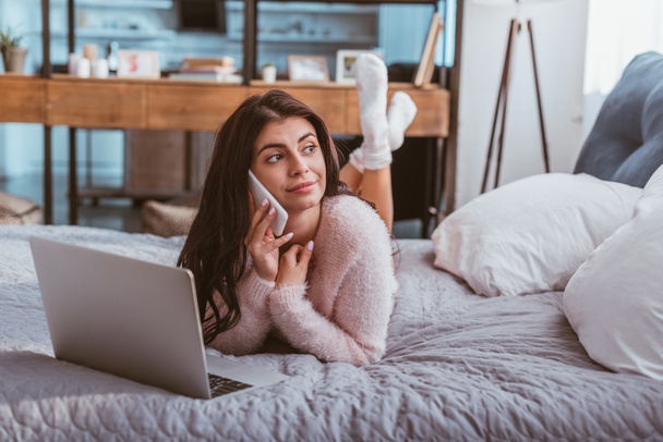 attractive young female freelancer talking on smartphone while laying on bed with laptop at home - Photo, Image
