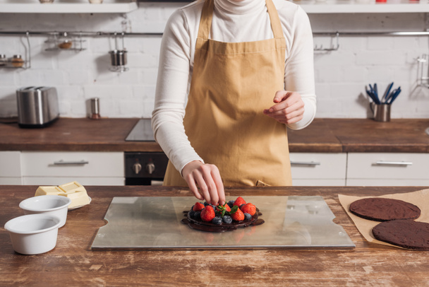 vista parcial de la mujer en delantal preparando delicioso pastel con bayas en la cocina
   - Foto, Imagen