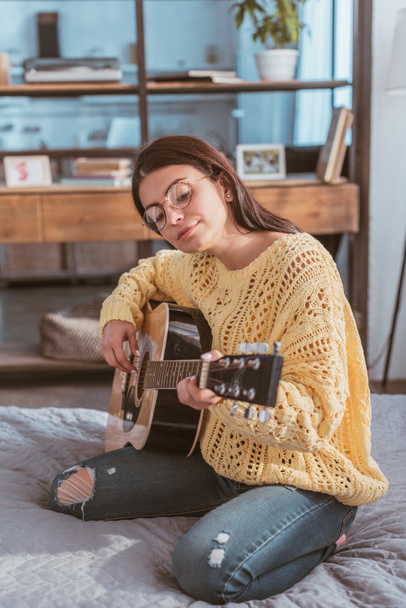 attractive woman in eyeglasses playing on acoustic guitar while sitting on bed at home - Foto, immagini