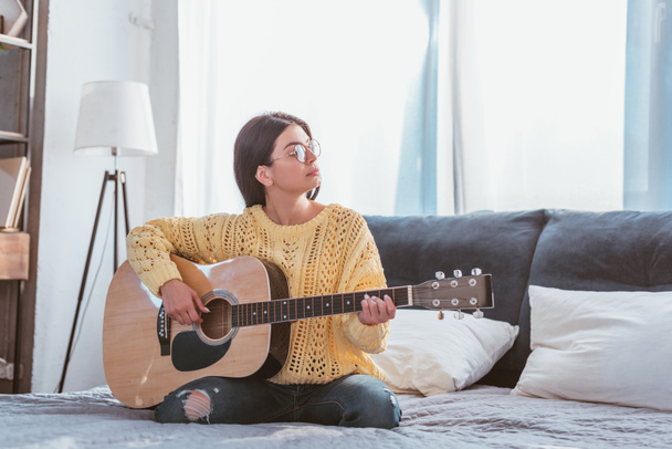young pretty woman in eyeglasses playing on acoustic guitar while sitting on bed at home - 写真・画像