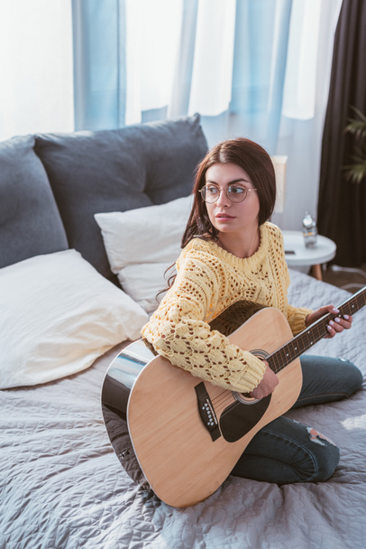vista de ángulo alto de la chica en gafas tocando en la guitarra acústica mientras está sentado en la cama en casa
 - Foto, imagen