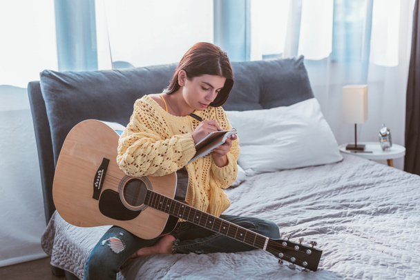 beautiful girl sitting with acoustic guitar and writing song in textbook on bed at home - Fotó, kép