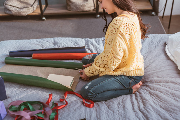 partial view of girl packing christmas gift while sitting on bed at home - Foto, Imagen