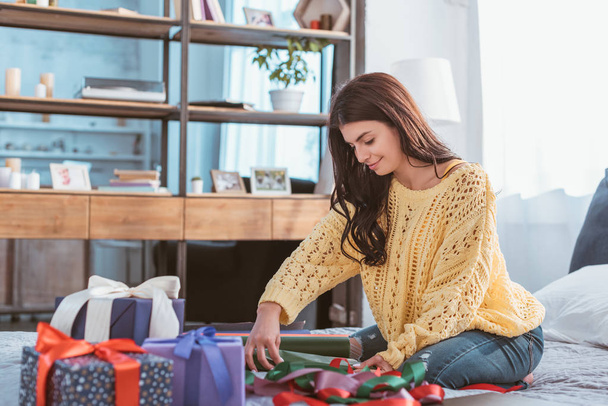 cheerful beautiful young woman packing gift while sitting on bed at home - Foto, afbeelding