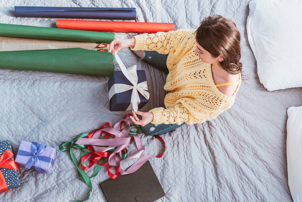Blick von oben auf junge Frau, die Geschenkschachtel mit festlichem Band umwickelt, während sie zu Hause auf dem Bett sitzt - Foto, Bild