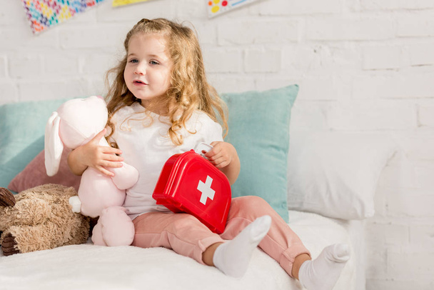 adorable kid holding first aid kit and rabbit toy in children room, looking away - Zdjęcie, obraz