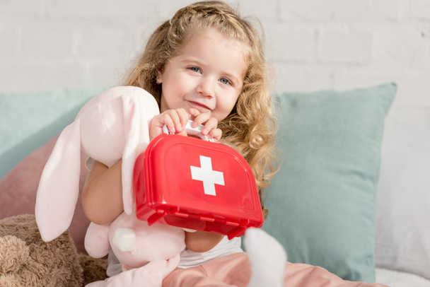 adorable kid holding first aid kit and rabbit toy in children room, looking at camera - Фото, изображение