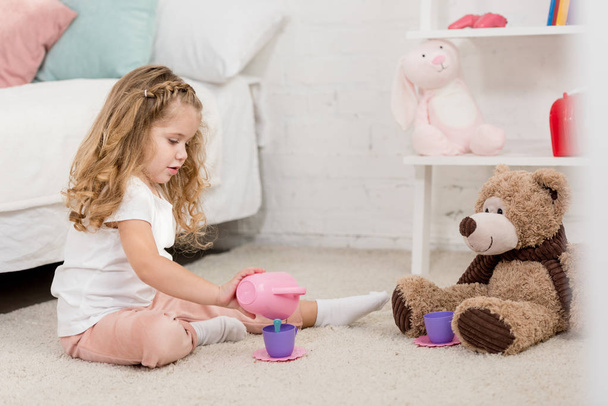 adorable kid playing with teddy bear and plastic cups on floor in children room - Foto, Bild