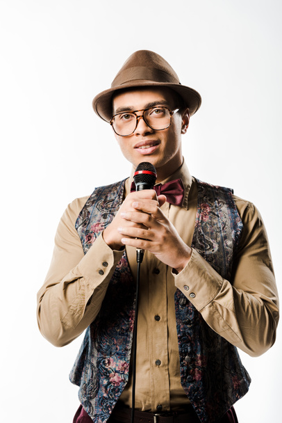 stylish mixed race man in hat and eyeglasses singing in microphone isolated on white - Photo, Image