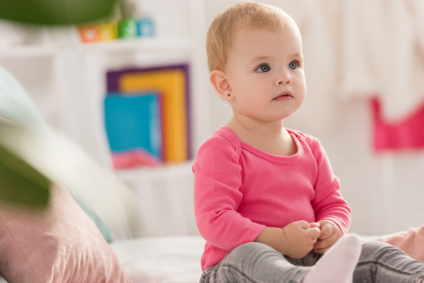 adorable niño en camisa rosa sentado en la cama y mirando hacia otro lado en la habitación de los niños
 - Foto, imagen