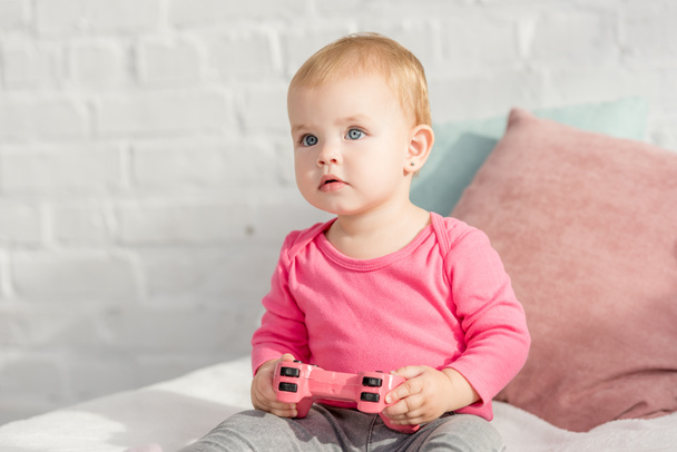 adorable kid in pink shirt holding pink joystick on bed in children room and looking up - Photo, Image
