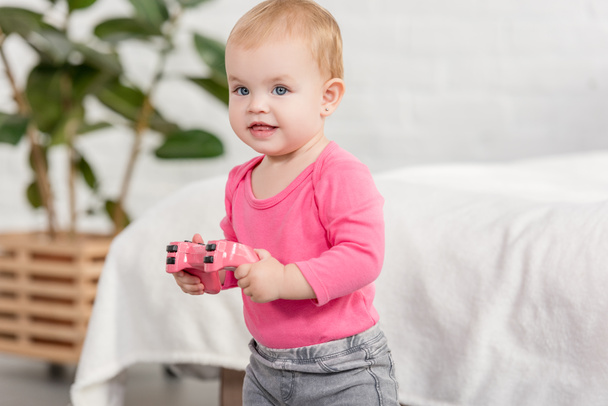 cheerful adorable kid in pink shirt holding pink joystick near bed in children room - Foto, Bild