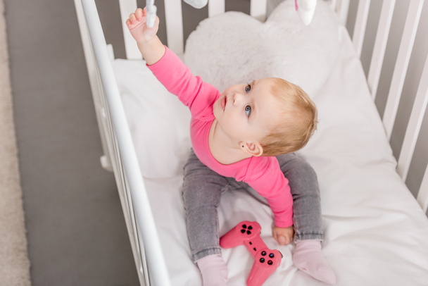 high angle view of adorable kid in pink shirt holding pink joystick in crib and reaching toy - Foto, afbeelding