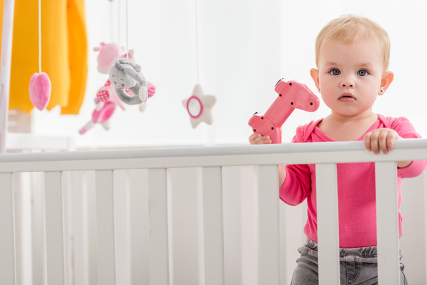 adorable kid in pink shirt holding pink joystick in crib and looking at camera - Fotografie, Obrázek