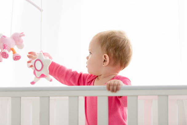 side view of adorable kid in pink shirt standing in crib and touching toys - Foto, Bild