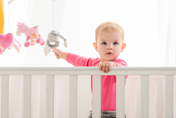adorable kid in pink shirt standing in crib and touching toys and looking up - Photo, Image