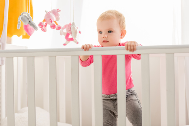 adorable toddler in pink shirt standing in crib and looking away - Valokuva, kuva
