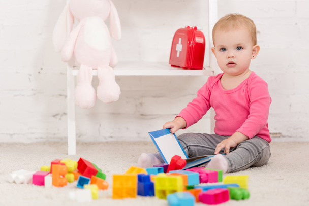 adorable kid sitting on carpet and holding book in children room - Foto, Imagen