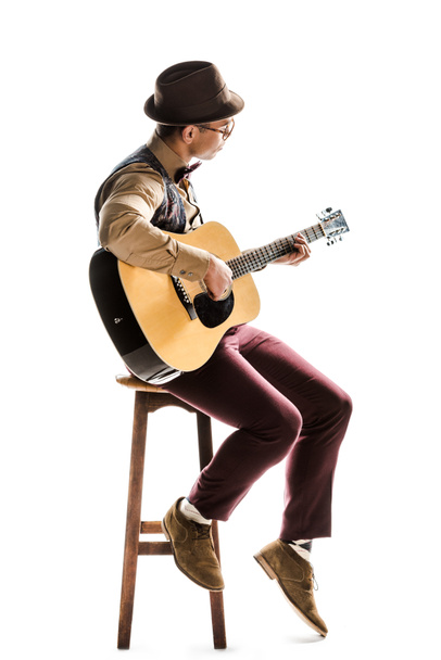 young mixed race male musician in hat and eyeglasses playing on acoustic guitar while sitting on chair isolated on white - 写真・画像