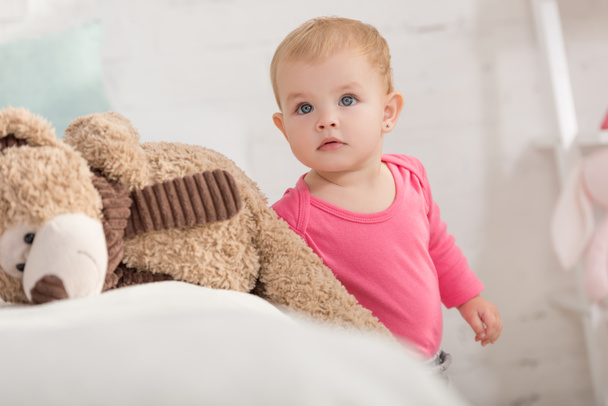 adorable kid standing near teddy bear and looking up in children room - Photo, Image