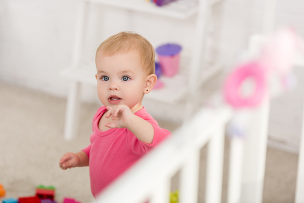 selective focus of adorable kid in pink shirt standing near crib in children room - Photo, Image