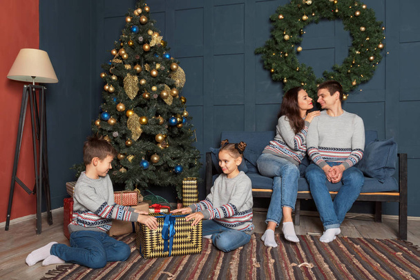 hermano y hermana están considerando una caja grande con un regalo cerca del árbol de Navidad en la habitación y los padres están sentados en el sofá
 - Foto, Imagen