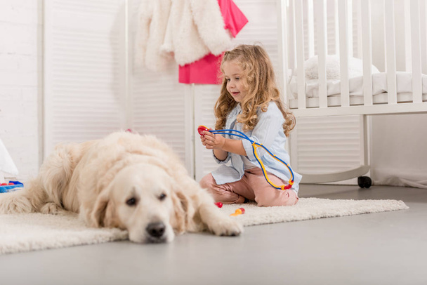 επιλεκτική εστίαση των αξιολάτρευτο παιδί παριστάνει κτηνίατρος και εξέταση golden retriever στο δωμάτιο παιδιών - Φωτογραφία, εικόνα
