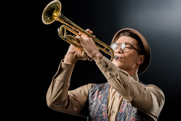 young male jazzman in hat and eyeglasses playing on trumpet isolated on black - Φωτογραφία, εικόνα