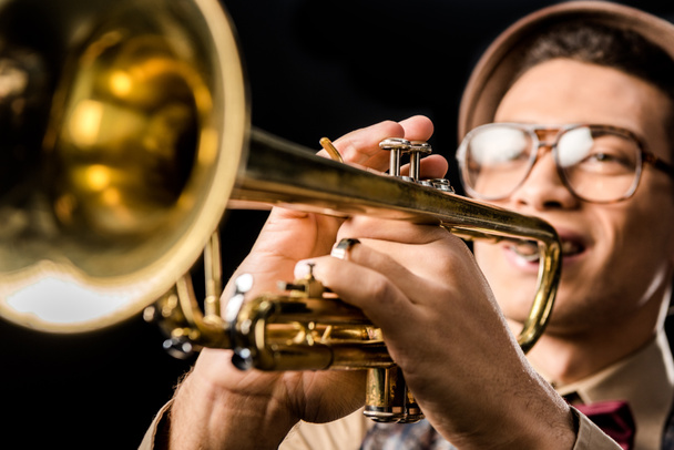 selective focus of male jazzman in hat and eyeglasses playing on trumpet isolated on black - Фото, изображение