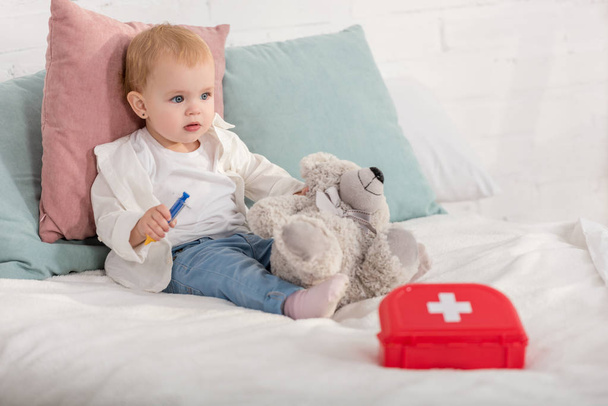 чарівна дитина сидить на ліжку з плюшевим ведмедем і аптечкою в дитячій кімнаті
 - Фото, зображення