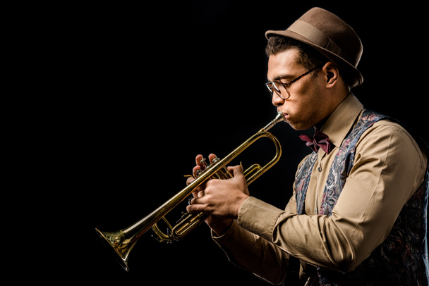 vista lateral da raça mista macho jazzman em chapéu e óculos tocando no trompete isolado no preto
 - Foto, Imagem