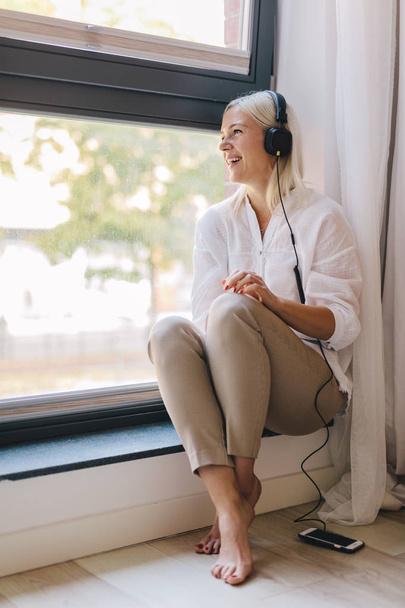 glückliche Frau, die Musik hört, auf einem Fensterbrett sitzend. Freizeit, Unterhaltung. - Foto, Bild