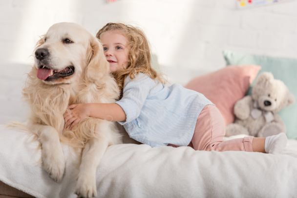 adorable kid hugging golden retriever dog on bed in children room - Photo, Image