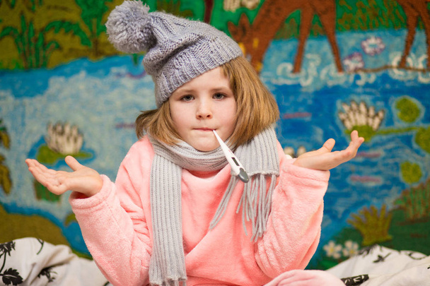 Хвора маленька дівчинка з сумним обличчям і термометром у роті носить шарф і капелюх
 - Фото, зображення